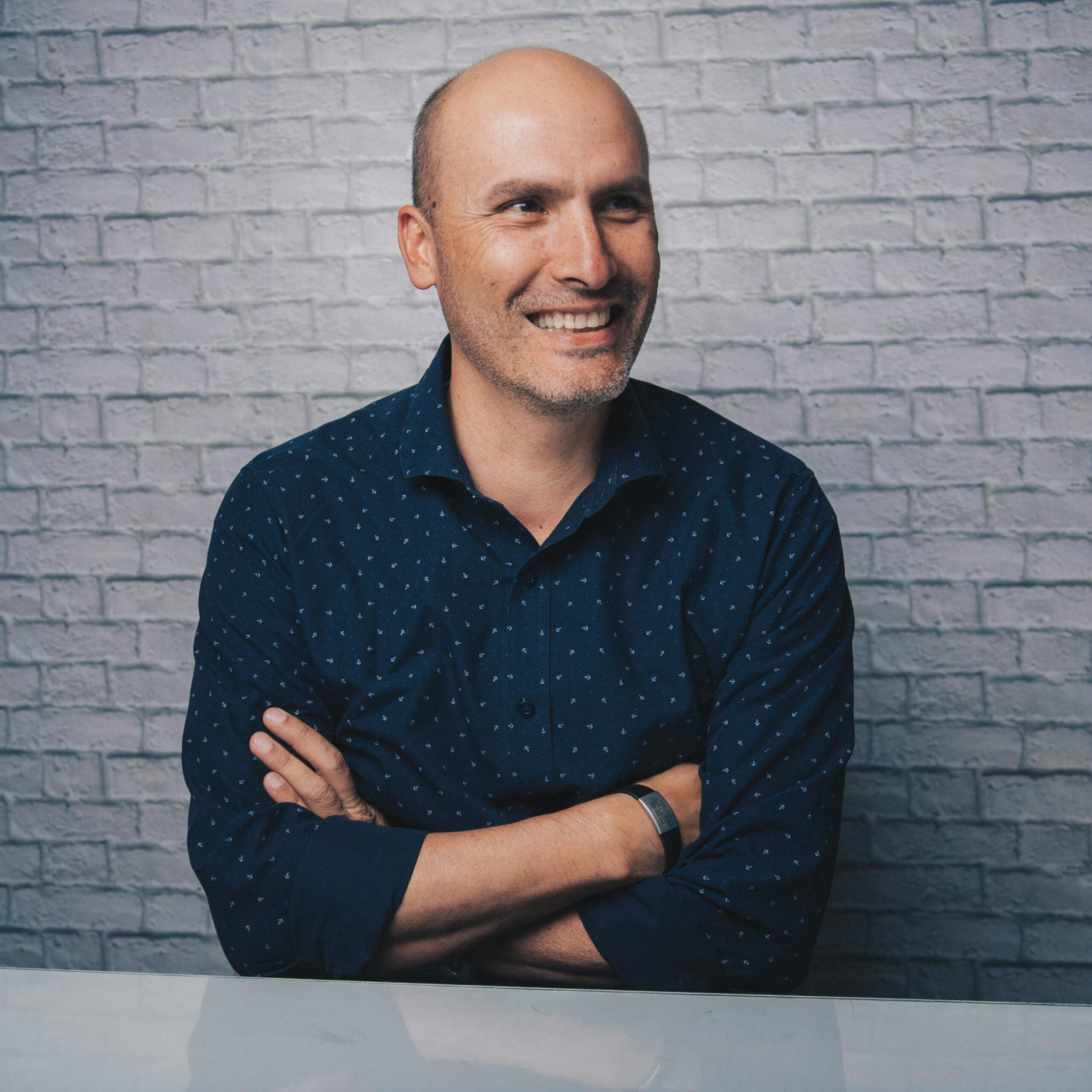 Sleep 001 Positive bald male in stylish shirt sitting at white table with crossed arms while looking away and smiling on white brick wall